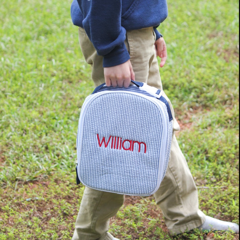 May include: A blue and white striped lunch bag with the name "William" embroidered in red. The bag has a handle and a white zipper. The person is wearing khaki pants and a navy blue sweatshirt.