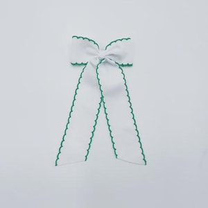 May include: A white hair bow featuring a central knot and two long, hanging tails. The edges of the bow and the tails are finished with a decorative green scalloped trim, providing a distinct contrast against the solid white fabric of the bow.