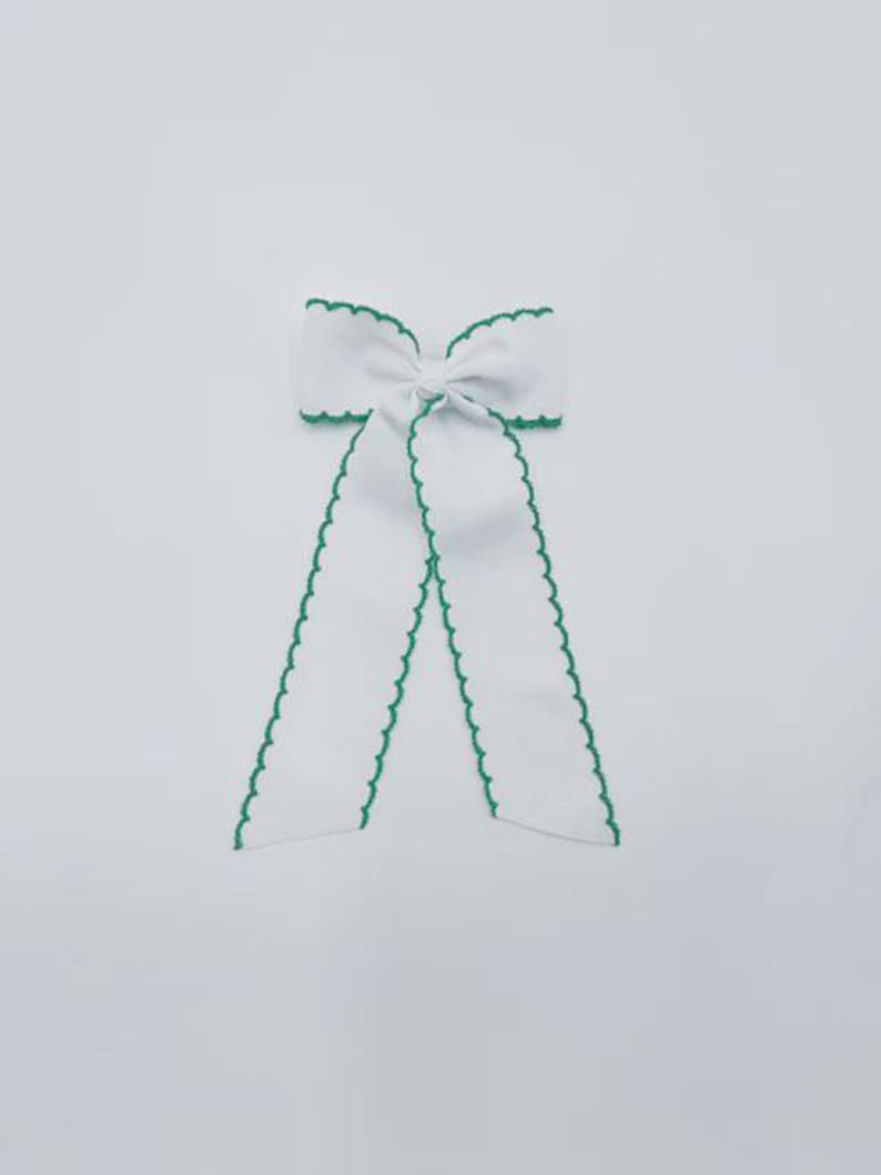 May include: A white hair bow featuring a central knot and two long, hanging tails. The edges of the bow and the tails are finished with a decorative green scalloped trim, providing a distinct contrast against the solid white fabric of the bow.