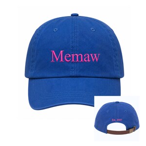 May include: A royal blue baseball cap with the embroidered word "Memaw" in pink across the front. The back of the cap features the text "Est. 2007" in pink. The cap has a brown adjustable strap.