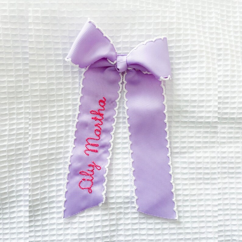 May include: A light purple ribbon hair bow featuring a white scalloped edge trim. The left tail of the bow is embroidered with the name "Lily Martha" in a bright pink cursive font. The ribbon has a ribbed texture throughout the entire design.