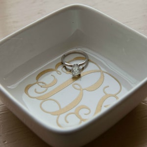 May include: A white ceramic ring dish with a gold monogram design. A silver ring with a clear stone is sitting in the dish.