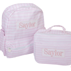 May include: A light pink and white gingham backpack and a matching rectangular lunch bag. Both items feature a white rectangular patch on the front with the name Saylor embroidered in pink thread. The bags have white zippers and trim.
