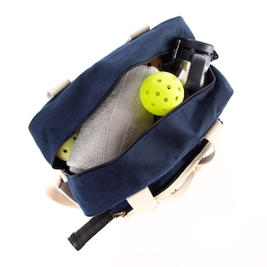 May include: A blue canvas duffel bag with white straps and a zippered top. The bag is open and contains a white towel, a silver water bottle, and two yellow pickleball balls.