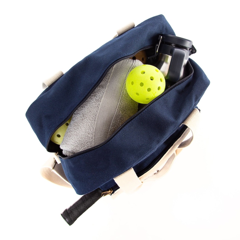 May include: A blue canvas duffel bag with white straps and a zippered top. The bag is open and contains a white towel, a silver water bottle, and two yellow pickleball balls.