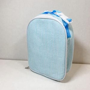 May include: A light blue and white striped lunch bag with a white trim. The bag has a top handle and a white buckle closure. The bag is approximately 9 inches tall, 8 inches wide, and 4 inches deep.