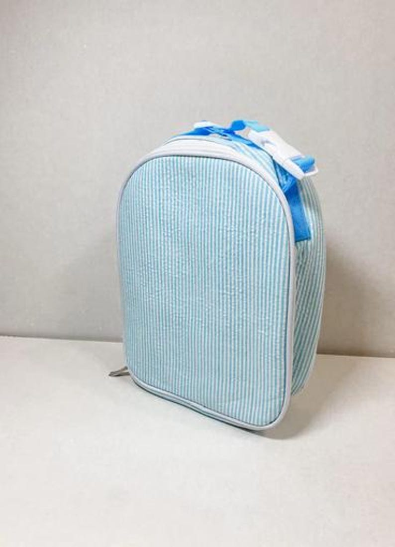 May include: A light blue and white striped lunch bag with a white trim. The bag has a top handle and a white buckle closure. The bag is approximately 9 inches tall, 8 inches wide, and 4 inches deep.