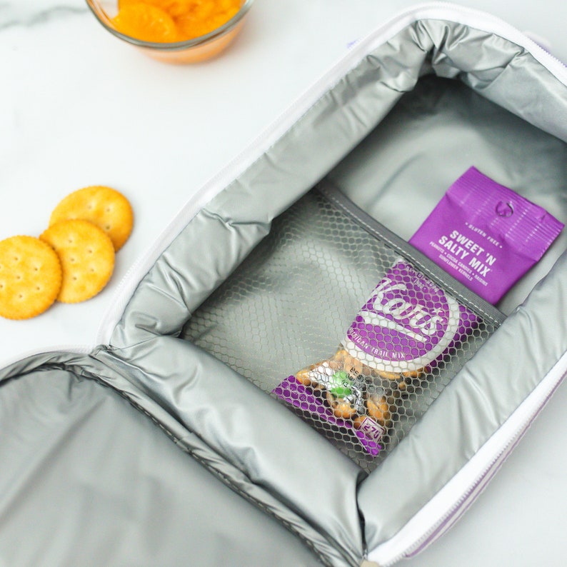May include: A white and purple insulated lunch bag is open, revealing a mesh pocket containing a bag of Kar's trail mix and a purple packet of Sweet 'N Salty Mix.  Round crackers and a bowl of orange snacks are visible.
