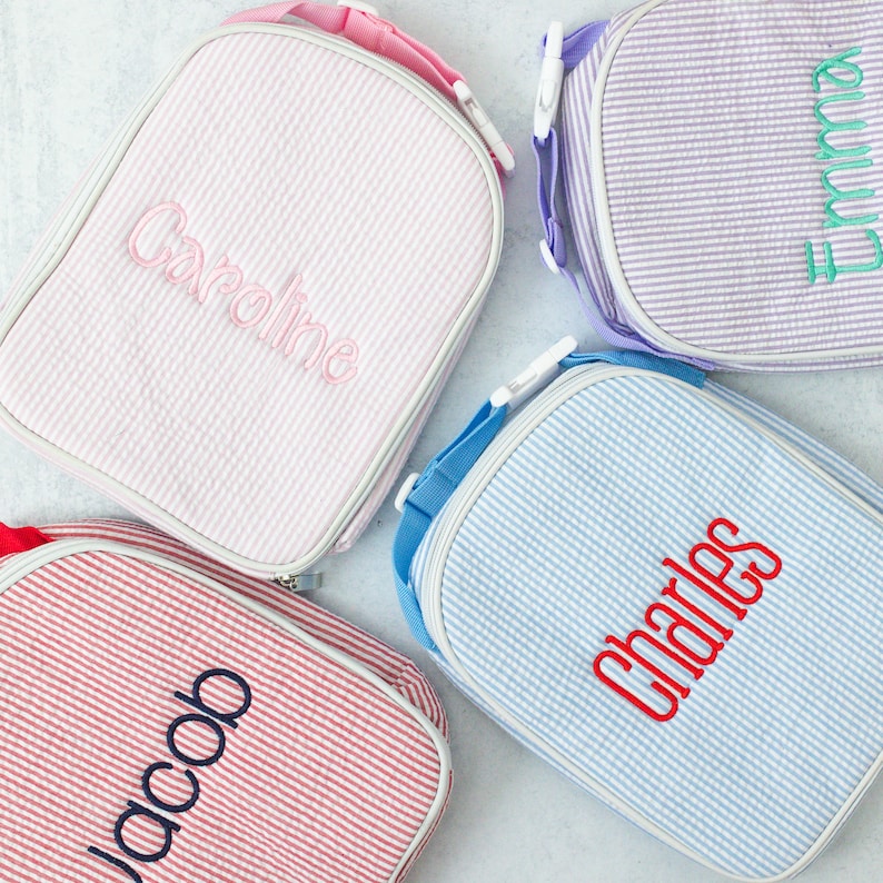 May include: Four personalized lunch bags in pastel colors: pink, purple, red, and blue. Each bag features a seersucker stripe pattern and a name embroidered in a contrasting color. The bags have a top handle and a shoulder strap.