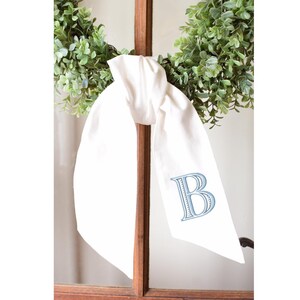 Op de afbeelding: Een wit stoffen lint geknoopt in een strik, voorzien van een geborduurde lichtblauwe letter B op een van de hangende uiteinden. Het lint is gedrapeerd over een groene bladerkrans die tegen een bruin houten raamkozijn is geplaatst.