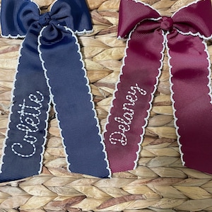 May include: Two grosgrain ribbon hair bows with scalloped white edges. The navy blue bow features the name Colette embroidered in white thread, and the burgundy bow features the name Delaney embroidered in white thread on the long tails.