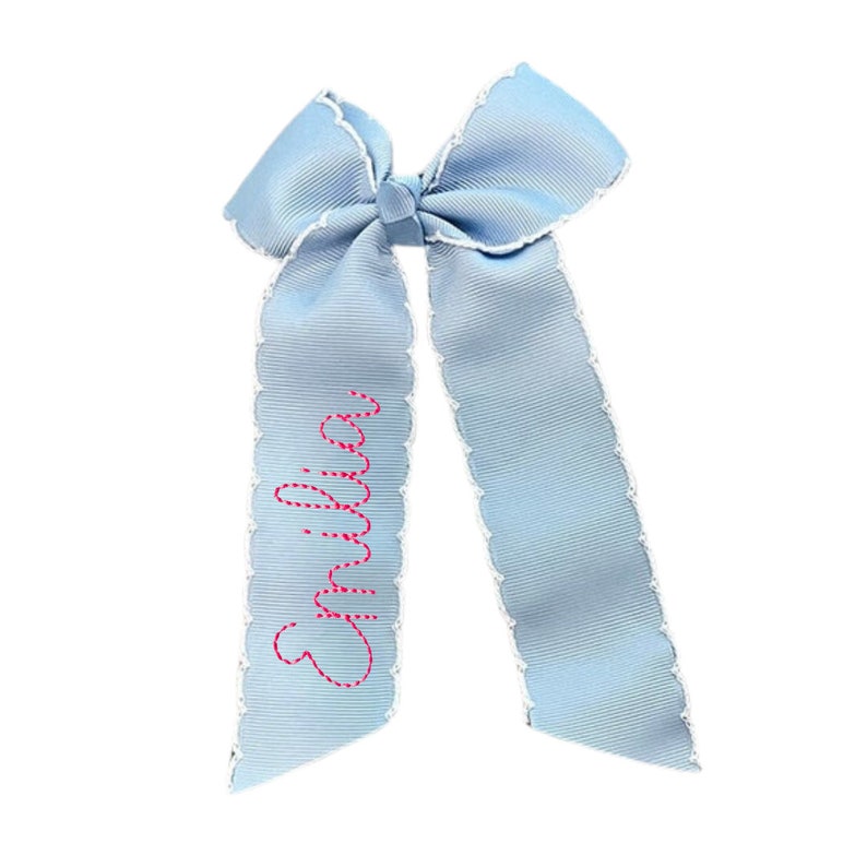 May include: A light blue hair bow made of ribbed fabric with a white scalloped edge trim. The left tail of the bow features the name Emilia embroidered in a bright pink cursive font. The bow has two long tails and a central knot detail.