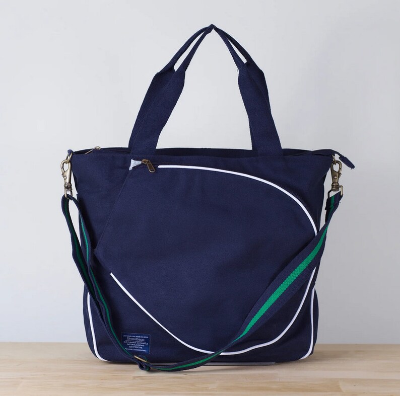 Puede incluir: Una bolsa de lona azul marino con un dise&ntilde;o de pelota de tenis blanca y ribete verde. La bolsa tiene un bolsillo con cremallera en la parte delantera y una correa de hombro larga ajustable.