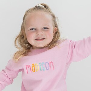 Puede incluir: Una sudadera rosa con el nombre "madison" bordado en colores arco&iacute;ris.