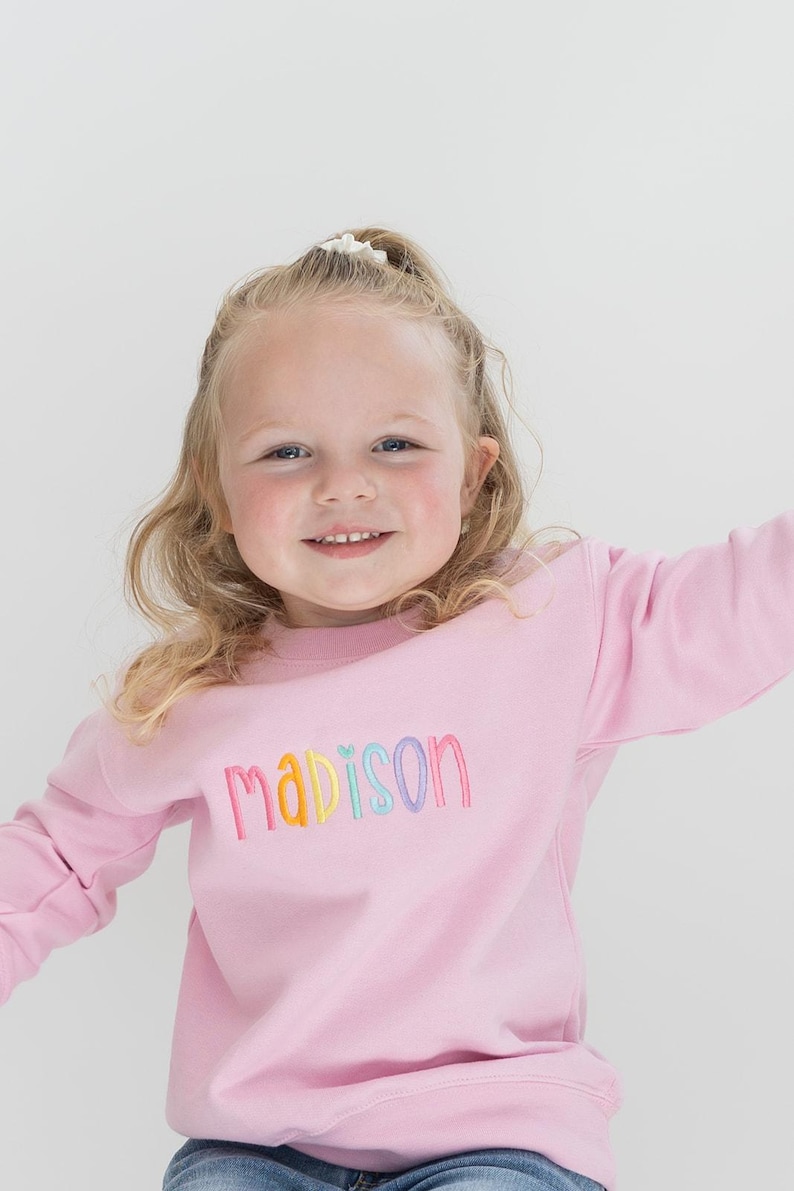Puede incluir: Una sudadera rosa con el nombre "madison" bordado en colores arco&iacute;ris.