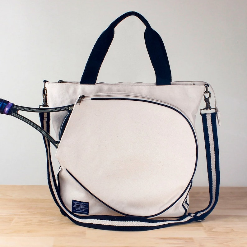 Puede incluir: Una bolsa de tenis de lona blanca con ribetes negros y una correa de hombro a rayas blancas y negras. La bolsa tiene un bolsillo integrado para raquetas y un bolsillo con cremallera en la parte delantera.