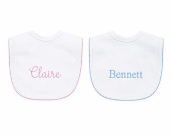 May include: Two white baby bibs with scalloped edges. The left bib features the name Claire embroidered in pink script, while the right bib features the name Bennett embroidered in blue block lettering. Both bibs have matching colored trim.