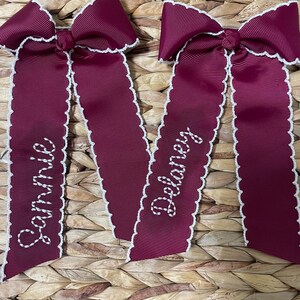 May include: Two deep burgundy hair bows made of ribbed fabric with white scalloped edges. Each bow features long, hanging tails with white embroidered names. One bow has the name Sammie stitched in cursive, and the other has Delaney.