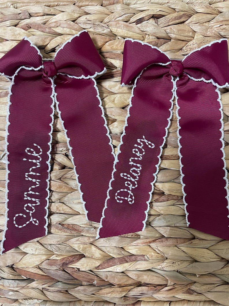 May include: Two deep burgundy hair bows made of ribbed fabric with white scalloped edges. Each bow features long, hanging tails with white embroidered names. One bow has the name Sammie stitched in cursive, and the other has Delaney.