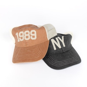 May include: Two baseball caps, one brown with a white "1989" embroidered on the front and a mesh back, and one black with a white "NY" embroidered on the front.