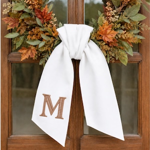 May include: A white fabric ribbon tied in a knot around an autumn wreath featuring green leaves, orange maple leaves, and small brown berries. The left tail of the ribbon features an embroidered brown letter M in a serif font style.