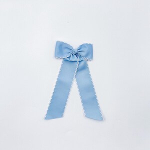 May include: A light blue hair bow made of ribbed fabric with a white scalloped edge trim. The bow features a central knot and two long, hanging ribbon tails that extend downwards, creating a symmetrical shape against a plain white background.