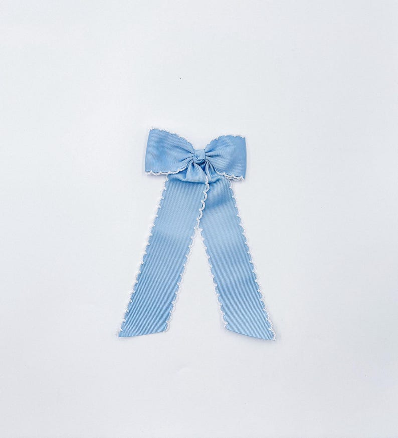 May include: A light blue hair bow made of ribbed fabric with a white scalloped edge trim. The bow features a central knot and two long, hanging ribbon tails that extend downwards, creating a symmetrical shape against a plain white background.