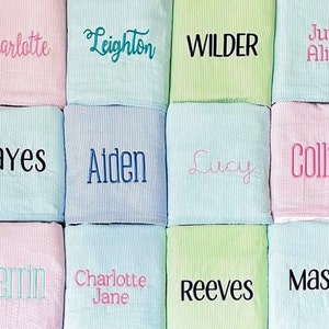 May include: Twelve personalized baby blankets in various colors, including pink, blue, and green. Each blanket features a different name embroidered in a contrasting color. The names include Charlotte, Leighton, Wilder, June Alice, Hayes, Aiden, Lucy, Collins, Merrin, Charlotte Jane, Reeves, and Mason.