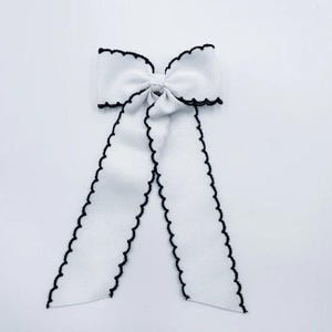 May include: A white fabric bow featuring a textured ribbon construction with a distinct black scalloped edge trim. The bow consists of a central knot with two long, hanging ribbon tails that extend downwards, each finished with the same black scalloped border.