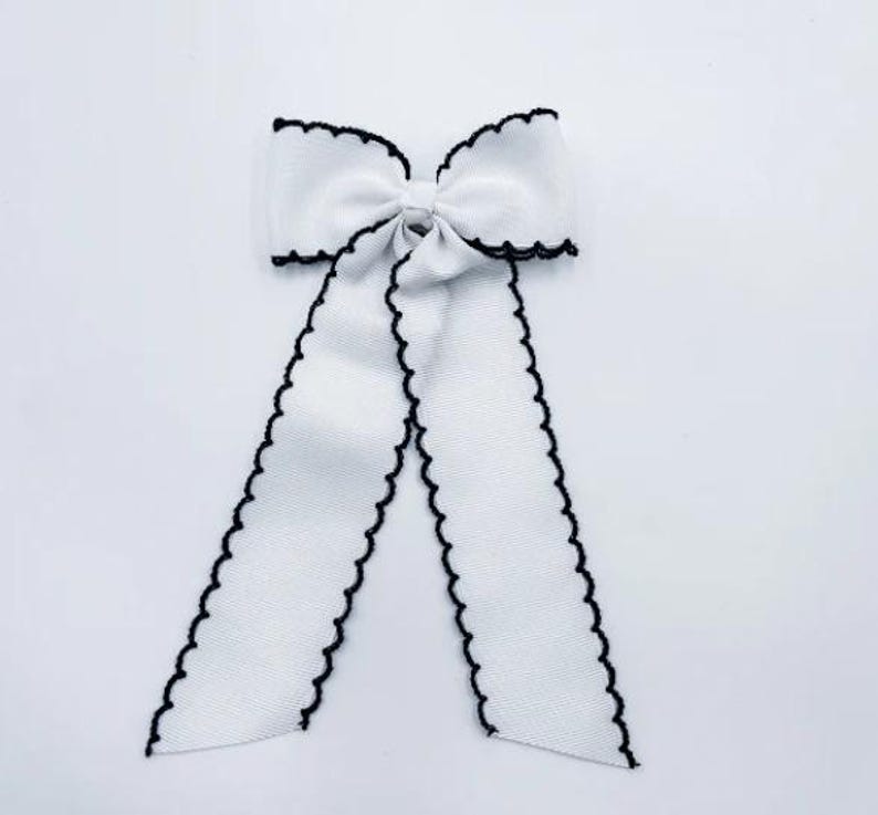 May include: A white fabric bow featuring a textured ribbon construction with a distinct black scalloped edge trim. The bow consists of a central knot with two long, hanging ribbon tails that extend downwards, each finished with the same black scalloped border.