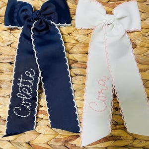 May include: Two hair bows made of grosgrain ribbon with scalloped edges. One bow is navy blue with white embroidery reading Colette, and the other is white with light pink embroidery reading Aria. Both bows feature long, hanging ribbon tails.