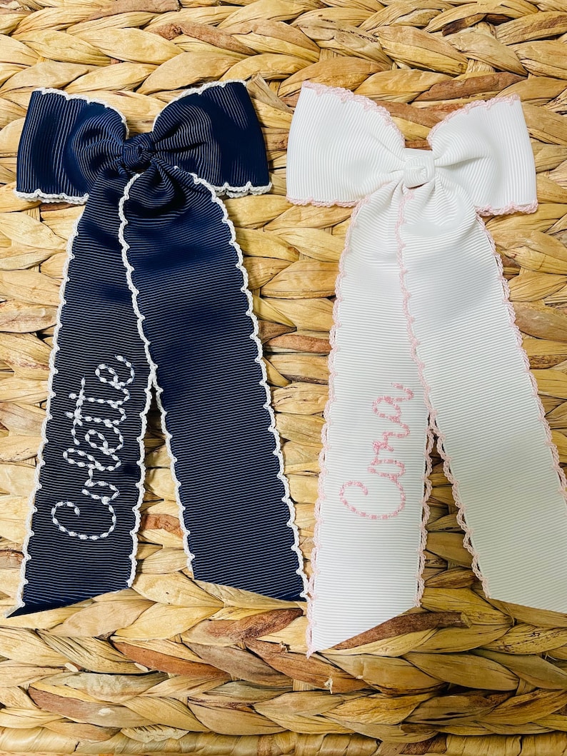 May include: Two hair bows made of grosgrain ribbon with scalloped edges. One bow is navy blue with white embroidery reading Colette, and the other is white with light pink embroidery reading Aria. Both bows feature long, hanging ribbon tails.