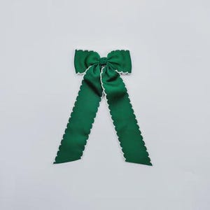 May include: A dark green fabric hair bow featuring a central knot and two long, downward-hanging tails. The edges of the bow and the tails are finished with a distinct white scalloped trim, providing a clear contrast against the solid green fabric.