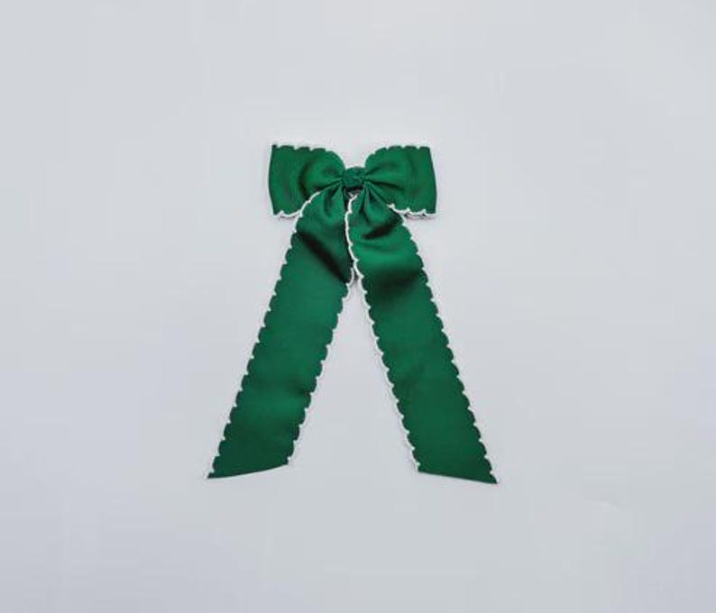 May include: A dark green fabric hair bow featuring a central knot and two long, downward-hanging tails. The edges of the bow and the tails are finished with a distinct white scalloped trim, providing a clear contrast against the solid green fabric.