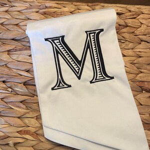Op de afbeelding: Een gevouwen off-white stoffen servet met een grote, centraal geplaatste, zwart geborduurde letter M. De letter is ontworpen in een vet schreeflettertype en bevat gedetailleerde interne lijnen binnen de structuur van het teken.
