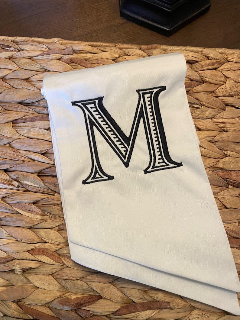 Op de afbeelding: Een gevouwen off-white stoffen servet met een grote, centraal geplaatste, zwart geborduurde letter M. De letter is ontworpen in een vet schreeflettertype en bevat gedetailleerde interne lijnen binnen de structuur van het teken.