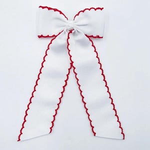 May include: A white hair bow made of ribbed fabric featuring a decorative red scalloped edge along the perimeter of the bow and its two long, hanging tails. The centre of the bow is knotted, creating a gathered appearance with symmetrical loops.
