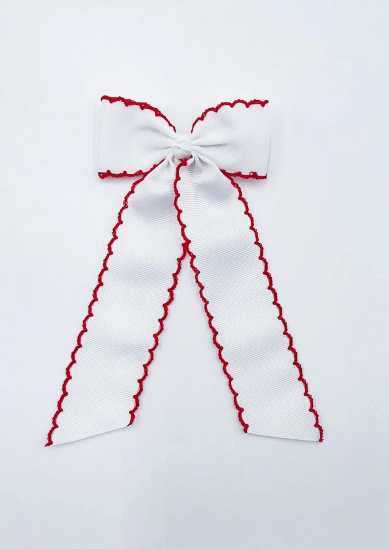 May include: A white hair bow made of ribbed fabric featuring a decorative red scalloped edge along the perimeter of the bow and its two long, hanging tails. The centre of the bow is knotted, creating a gathered appearance with symmetrical loops.