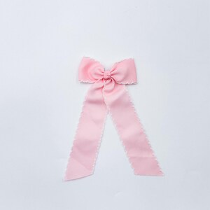 May include: A light pink ribbon bow featuring a central knot and two long, downward-hanging tails. The edges of the ribbon are finished with a delicate white scalloped trim, providing a subtle contrast to the solid pink fabric texture.