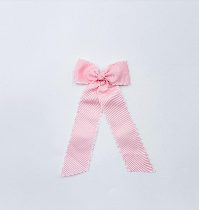 May include: A light pink ribbon bow featuring a central knot and two long, downward-hanging tails. The edges of the ribbon are finished with a delicate white scalloped trim, providing a subtle contrast to the solid pink fabric texture.