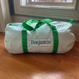 May include: A green and white striped duffel bag with green handles and trim. The name "Benjamin" is embroidered in green on a white rectangular patch on the front of the bag. The bag is sitting on a wooden floor.