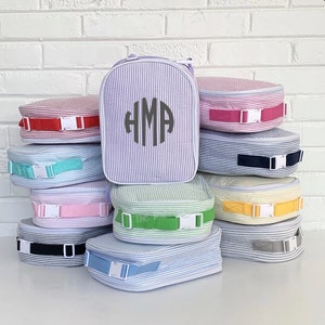 May include: A stack of seersucker lunch bags and a matching backpack. The bags and backpack are white with vertical stripes in various colors, including red, pink, blue, green, and yellow. The backpack has a monogram and a handle.