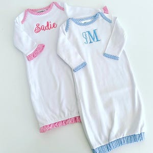 May include: Two white baby gowns with gingham trim. One gown has pink gingham trim and is embroidered with the name "Sadie". The other gown has blue gingham trim and is embroidered with the monogram "JM".