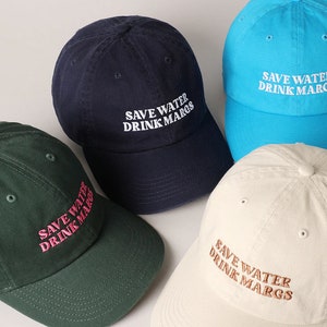 May include: Four baseball caps in different colors with the text "Save Water Drink Margs" embroidered on the front. The caps are navy blue, turquoise, green, and beige.