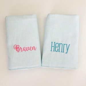 May include: Two light blue baby blankets with white stripes. One blanket has the name "Braven" embroidered in pink. The other blanket has the name "Henry" embroidered in teal.