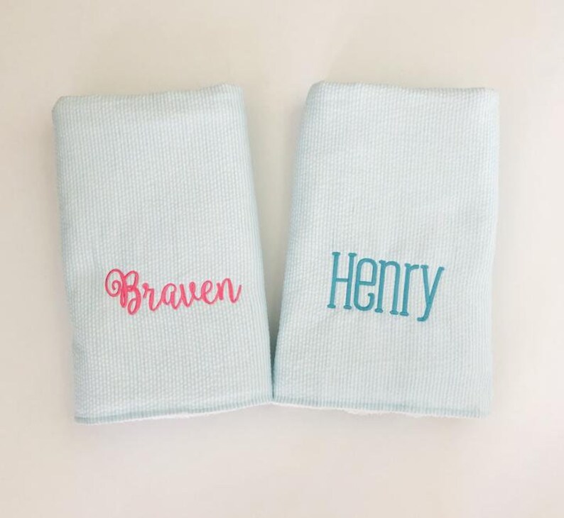 May include: Two light blue baby blankets with white stripes. One blanket has the name "Braven" embroidered in pink. The other blanket has the name "Henry" embroidered in teal.