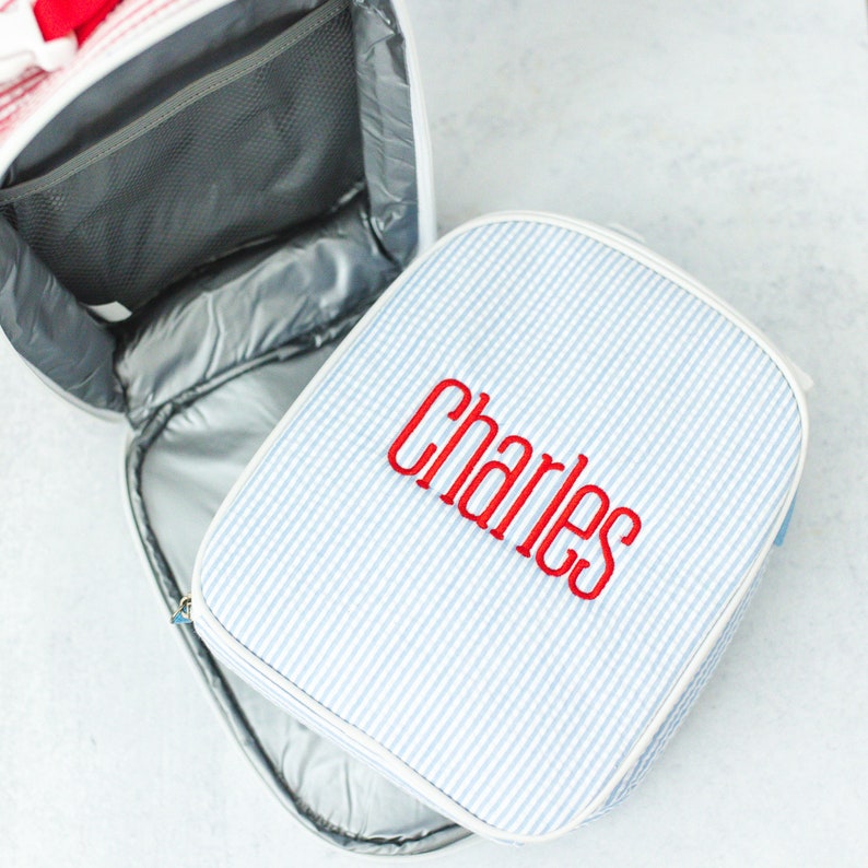 May include: A light blue and white striped lunch bag with the name "Charles" embroidered in red. The bag is open, revealing a silver insulated interior and a mesh pocket. A second, matching lunch bag is also visible.