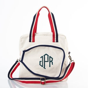 May include: A white canvas tote bag with a navy blue trim and red and navy blue straps. The bag has a monogram of the letters "JPR" in a dark green color.