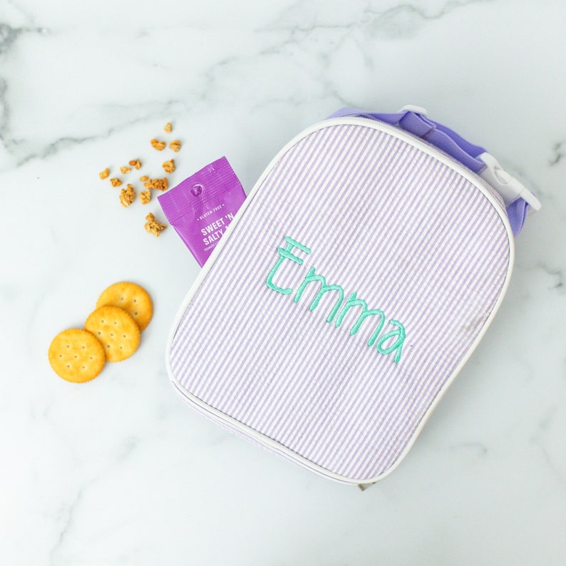 May include: A light purple and white striped lunch bag with the name "Emma" embroidered in teal. The bag is open, with a purple snack packet and crackers visible. The bag has a white trim and a purple handle.