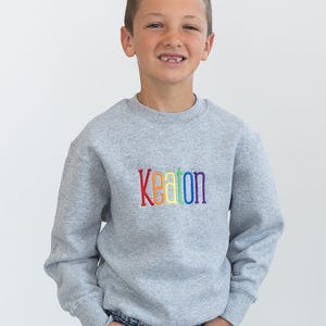 Puede incluir: Una sudadera gris con el nombre "Keaton" bordado en arco&iacute;ris en la parte delantera. La sudadera la lleva puesta un joven que lleva unos vaqueros azules.